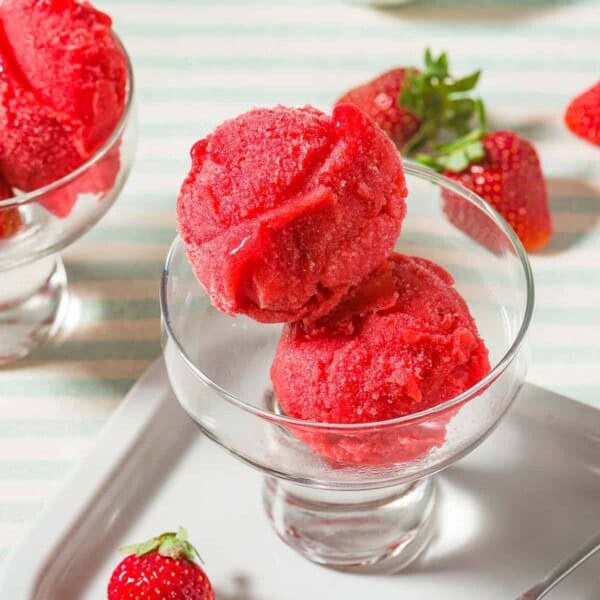 Two scoops of strawberry sorbet in a bowl next to a spoon, several scattered strawberries, a small bowl of strawberries and another bowl of the sorbet.