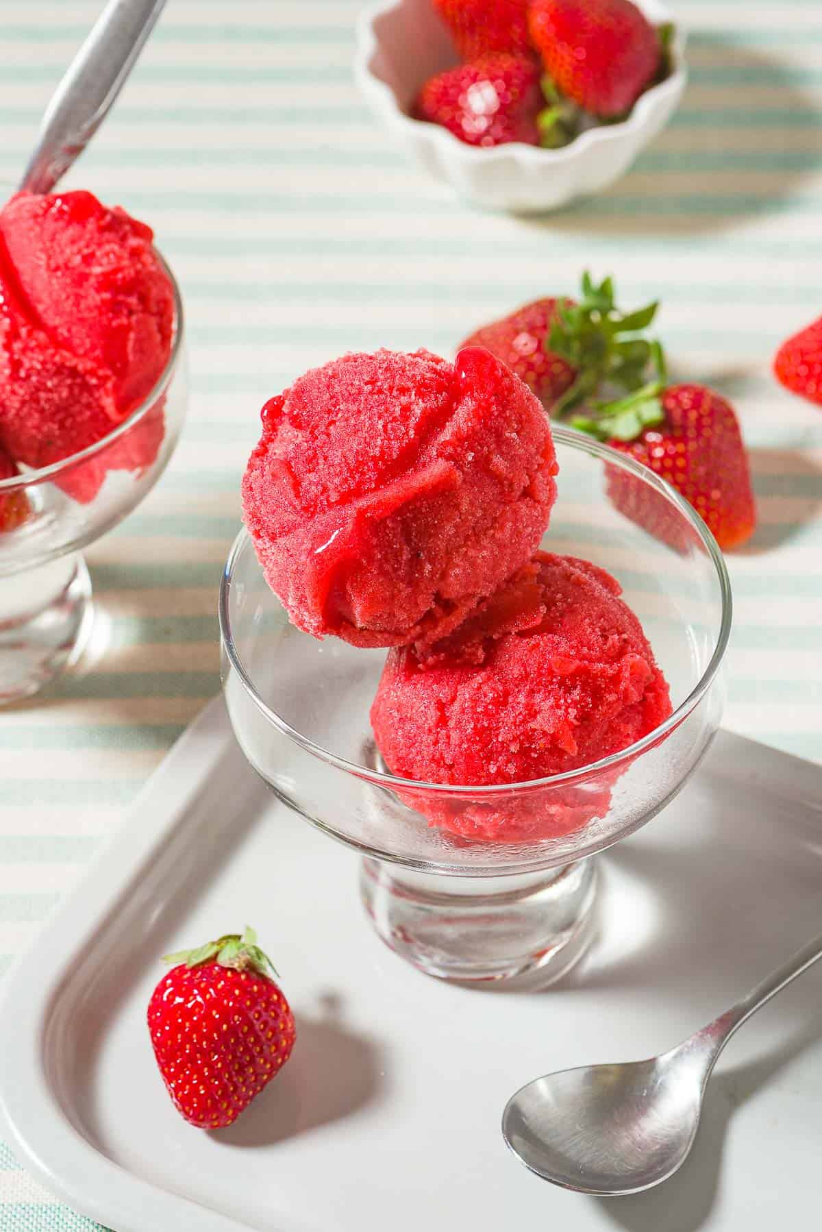 Two scoops of strawberry sorbet in a bowl next to a spoon, several scattered strawberries, a small bowl of strawberries and another bowl of the sorbet.