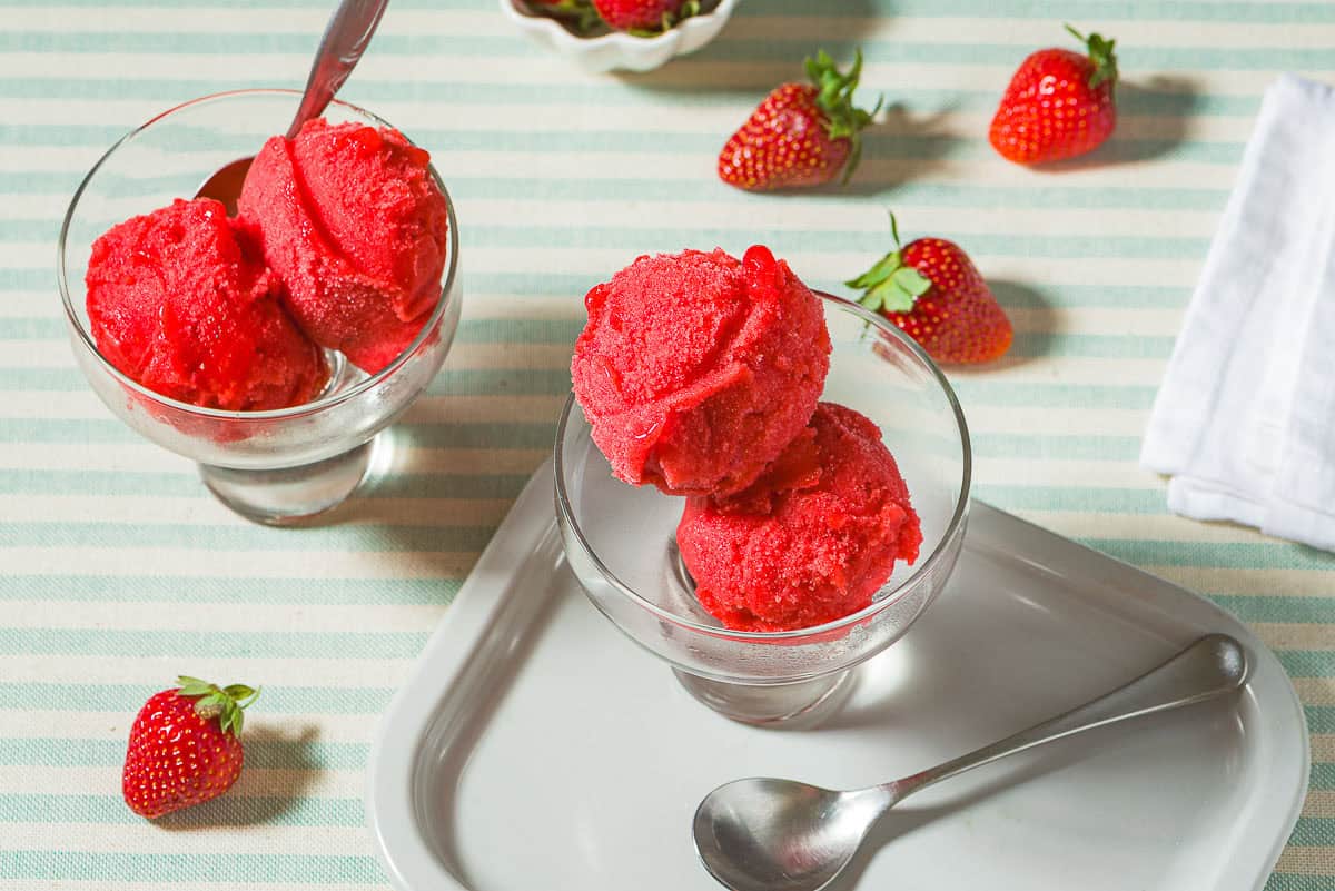 Two bowls of strawberry sorbet, one with a spoon. Next to these is a spoon, a small bowl of strawberries, several scattered strawberries, and a cloth napkin.