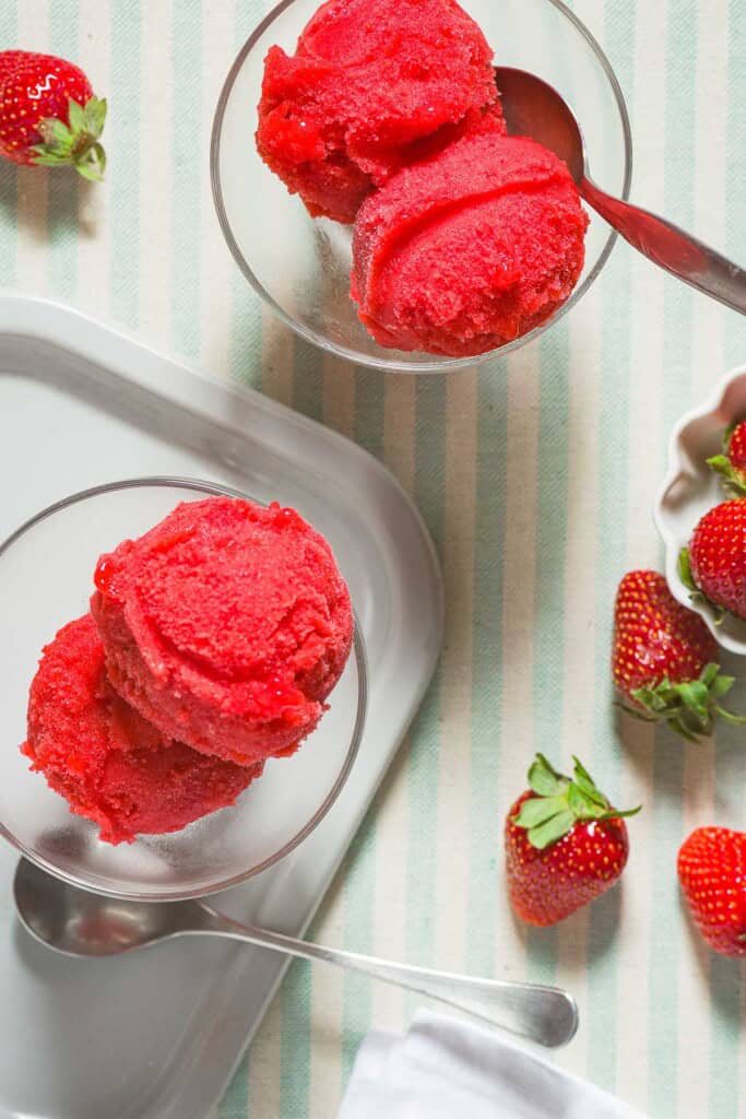 Two bowls of strawberry sorbet, one with a spoon. Next to these is a spoon, a small bowl of strawberries, and several scattered strawberries.