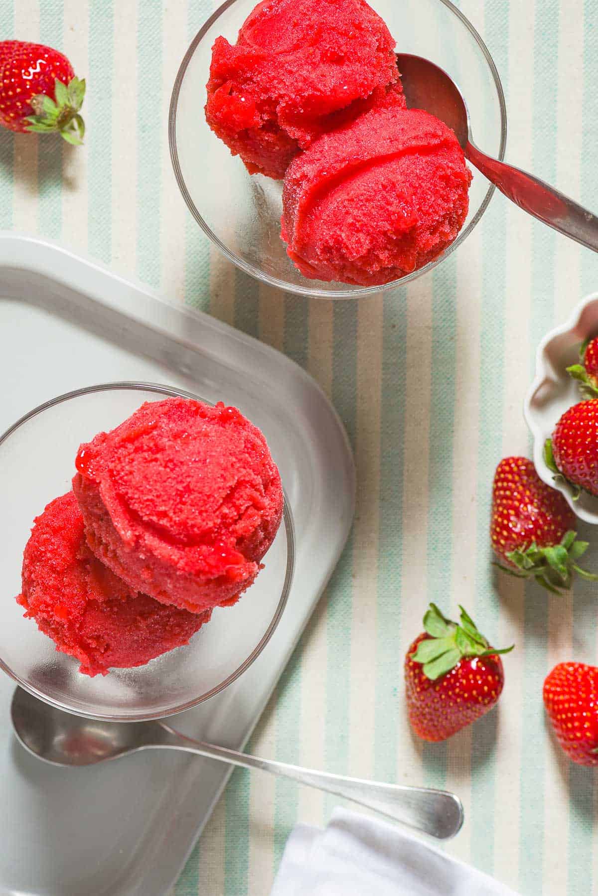 Two bowls of strawberry sorbet, one with a spoon. Next to these is a spoon, a small bowl of strawberries, and several scattered strawberries.