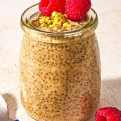 A jar of chia seed pudding topped with raspberries and crushed pistachios next to a spoon.