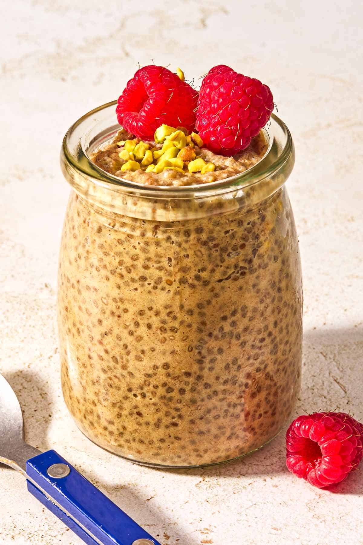 A jar of chia seed pudding topped with raspberries and crushed pistachios next to a spoon.