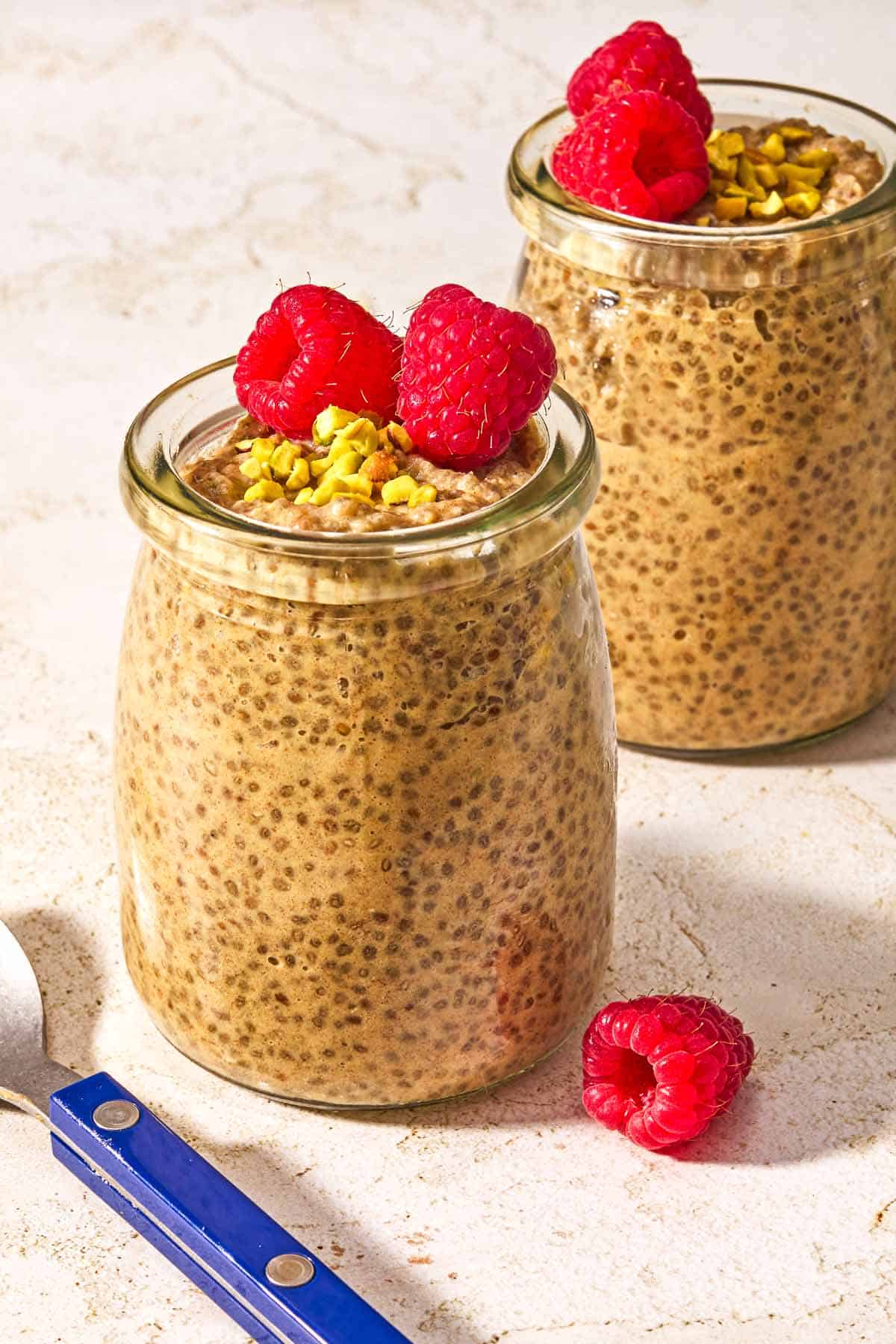 Two jars of chia seed pudding topped with raspberries and crushed pistachios next to a spoon.