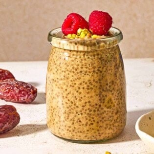 A jar of chia seed pudding topped with raspberries and crushed pistachios next to three whole dates and a bowl of crushed pistachios.