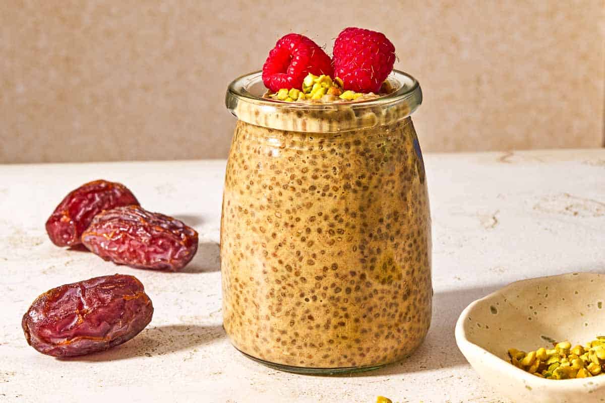A jar of chia seed pudding topped with raspberries and crushed pistachios next to three whole dates and a bowl of crushed pistachios.