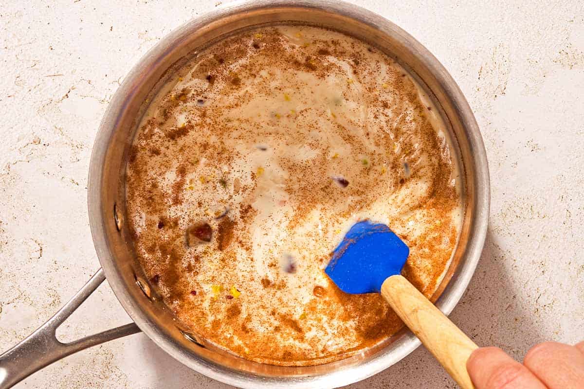 The milk, dates, pistachios cardamom and cinnamon being stirred together in a saucepan with a spatula.