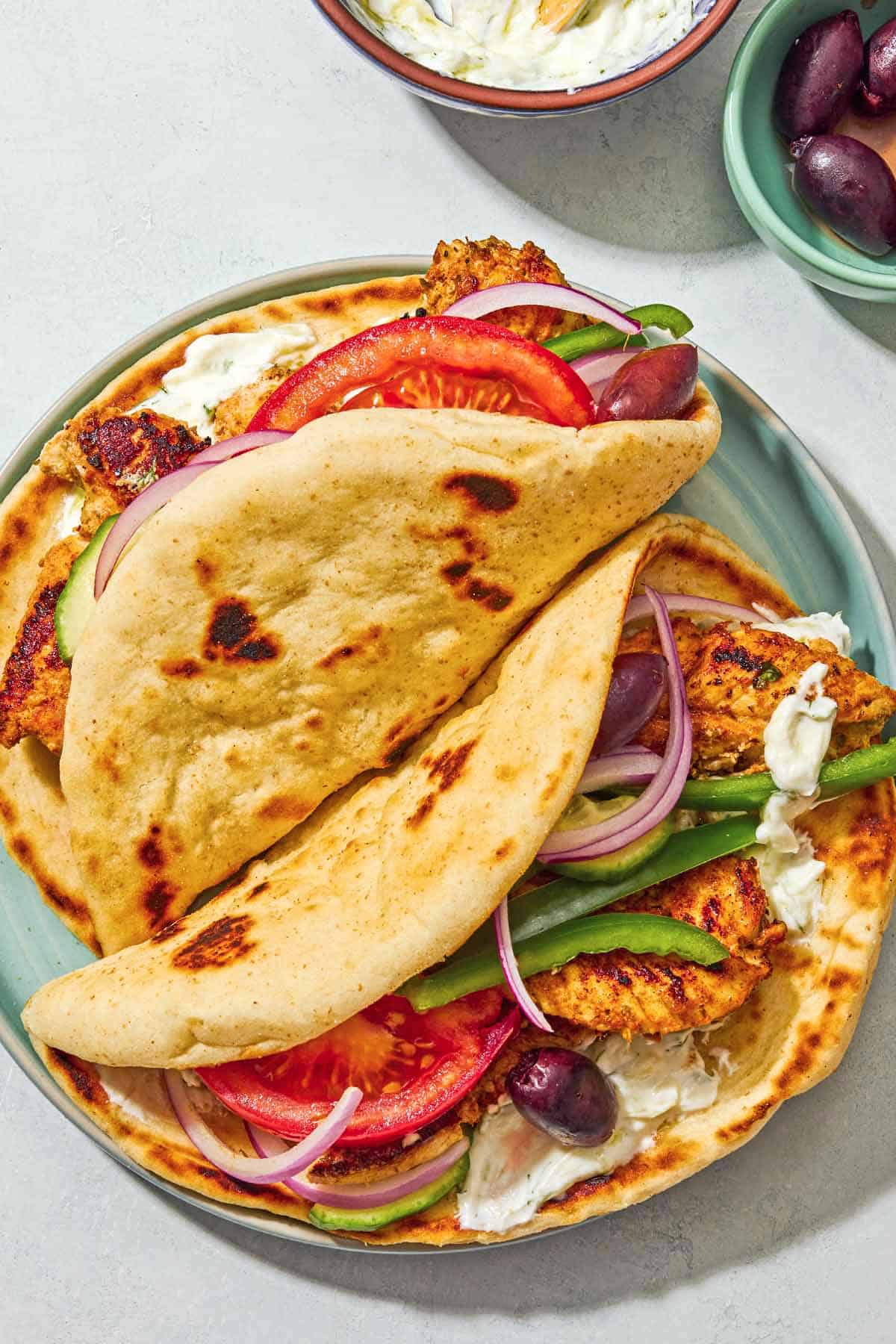 Two chicken gyros on a plate next to bowls of tzatziki sauce and kalamata olives.