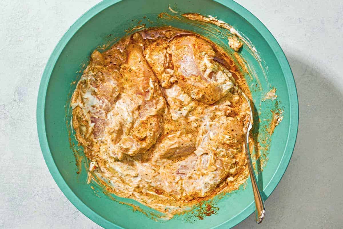 The chicken tenders for the chicken gyro recipe in a bowl of marinade with a spoon.