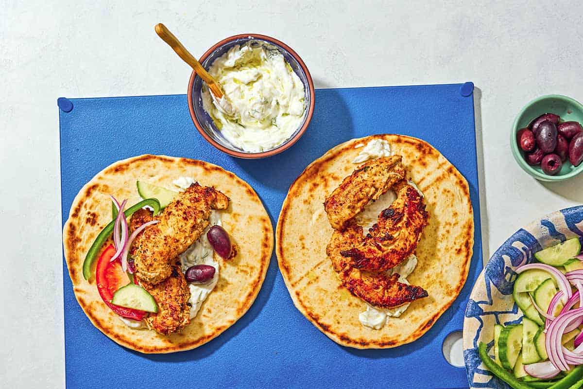 Two chicken gyros being assembled on a cutting board with a bowl of tzatziki. Next to this is a small bowl of kalamata olives and a bowl of sliced cucumbers, red onion and green pepper.