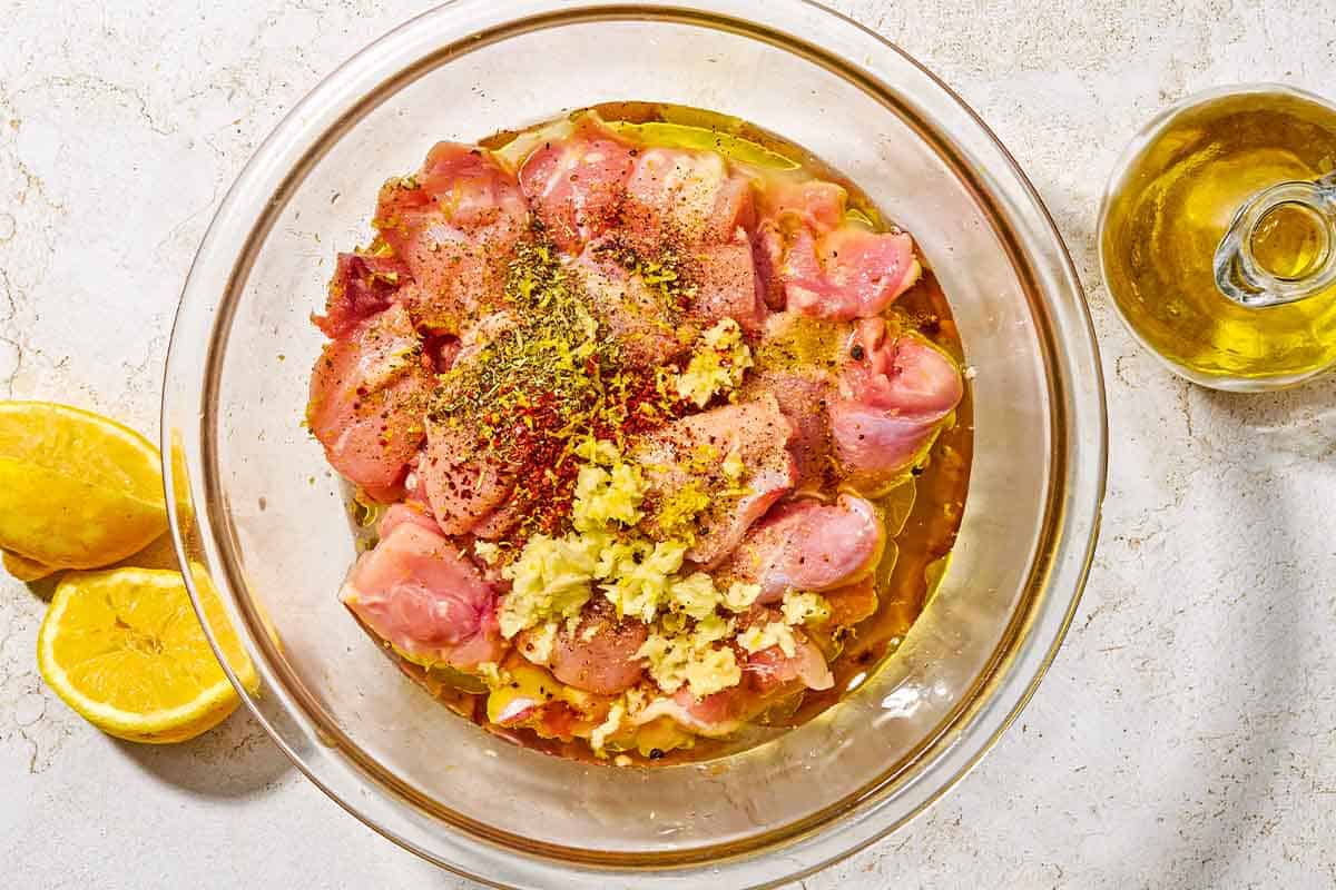 Cubed chicken thigh pieces in a bowl with the marinade ingredients just before being fully mixed together. Next to this is a bottle of olive oil and 2 juiced lemon halves.