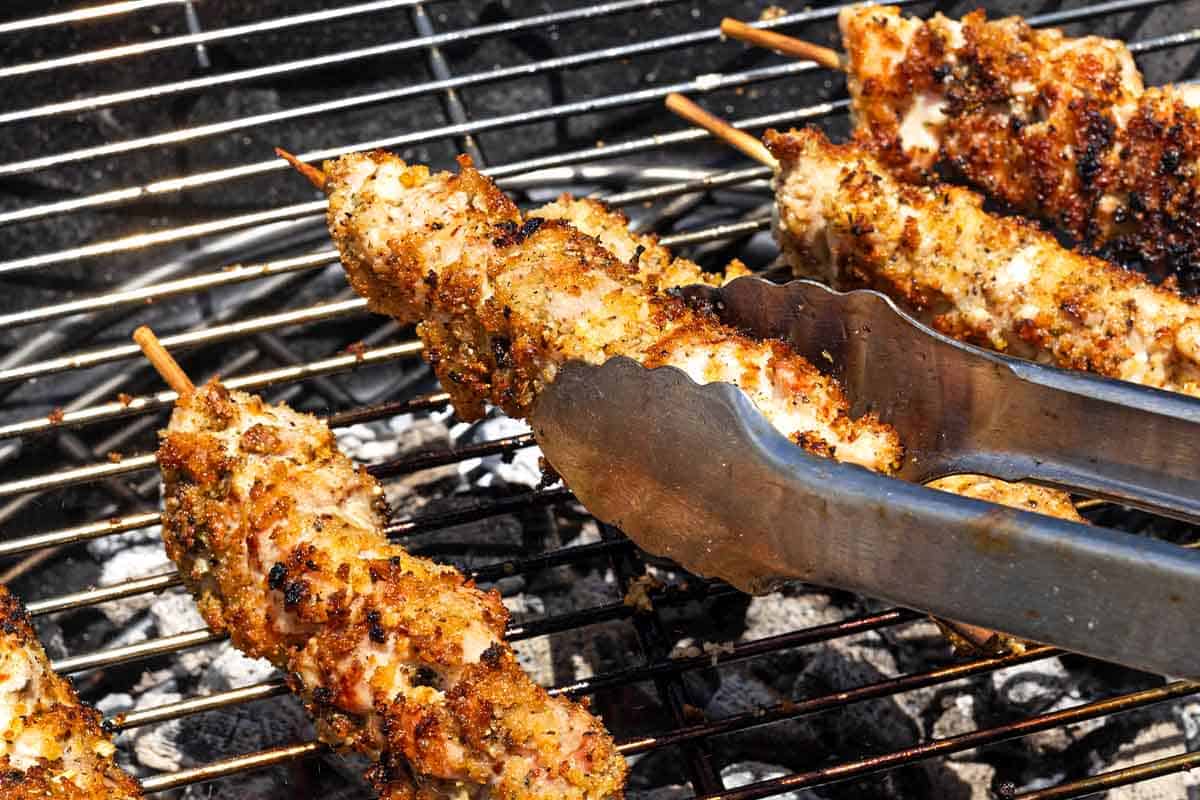 A chicken spiedini skewer being turned on a grill with a pair of tongs next to four more skewers.