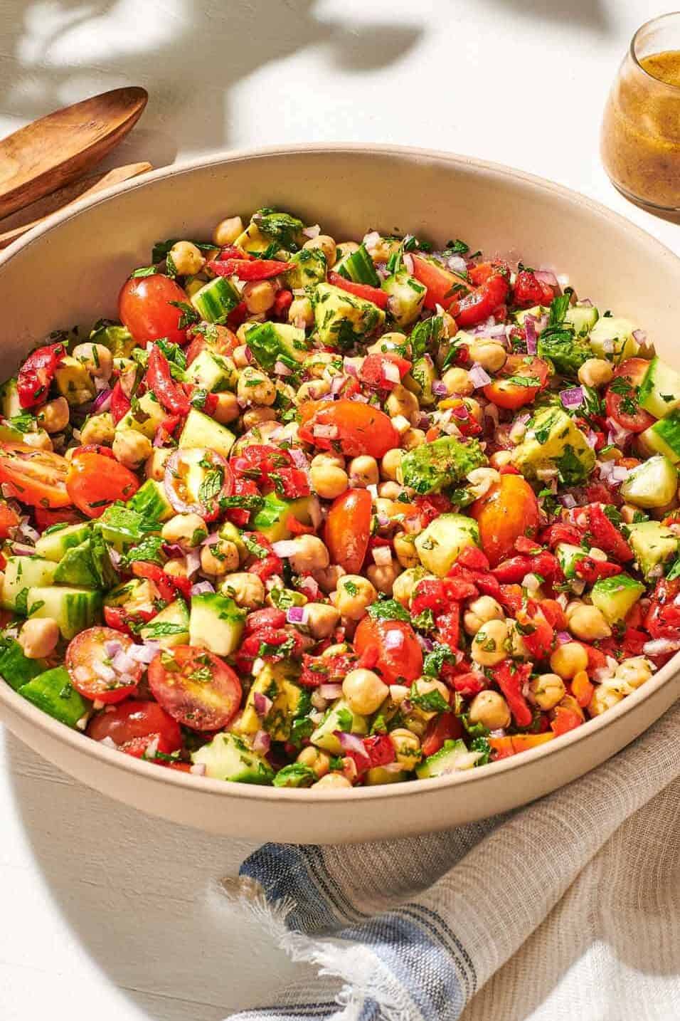 a close up of vegan chickpea salad in a serving bowl next to a wooden serving spoon and fork, a jar of dijon dressing, and a cloth napkin.