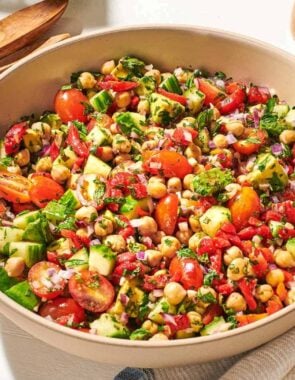 a close up of vegan chickpea salad in a serving bowl next to a wooden serving spoon and fork, a jar of dijon dressing, and a cloth napkin.