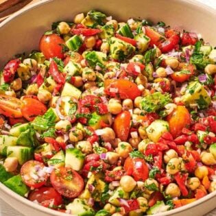 a close up of vegan chickpea salad in a serving bowl next to a wooden serving spoon and fork, a jar of dijon dressing, and a cloth napkin.