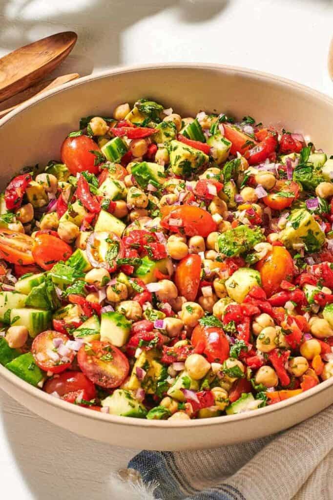 a close up of vegan chickpea salad in a serving bowl next to a wooden serving spoon and fork, a jar of dijon dressing, and a cloth napkin.