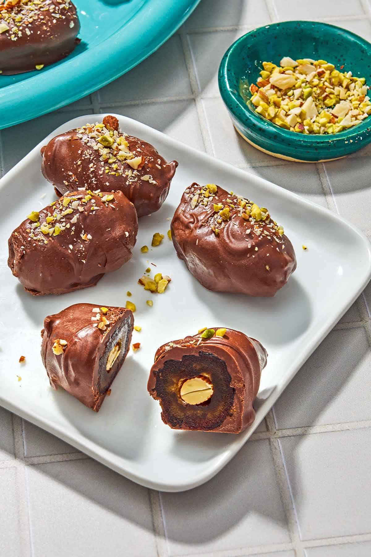 4 chocolate covered dates on a plate, on cut in half to show an almond inside. Next to this is a small bowl of crushed pistachios and almonds.