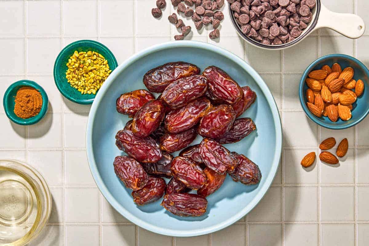 Ingredients for chocolate covered dates including dates, chocolate chips, roasted almonds, crushed pistachios, olive oil, and cinnamon.