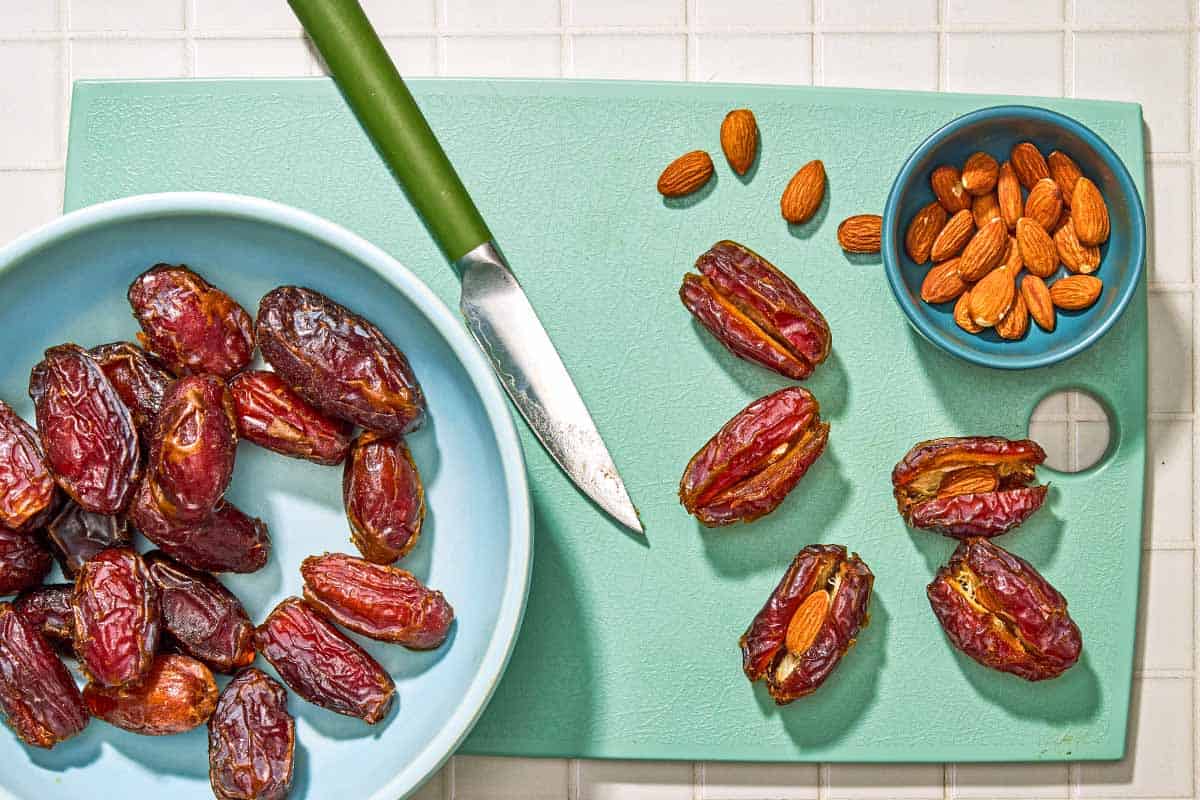 A bowl of dates on a cutting board with a knife, a bowl of toasted almonds and 5 dates slit open, 2 with almonds placed inside.