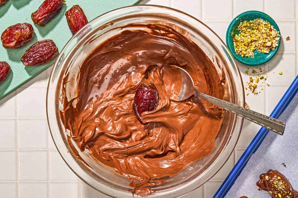 A date in a bowl of chocolate with a spoon next to a cutting board with dates, a bowl of crushed almonds and pistachios and a parchment lined sheet pan with a chocolate covered almond.