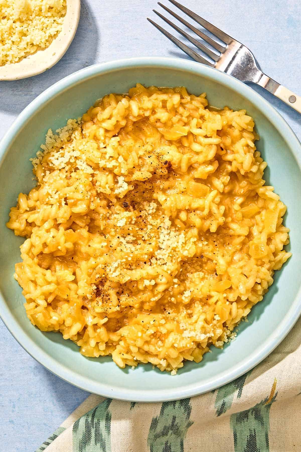 Risotto in a bowl next to a fork, a cloth napkin and a small bowl of Parmigiano-Reggiano cheese.