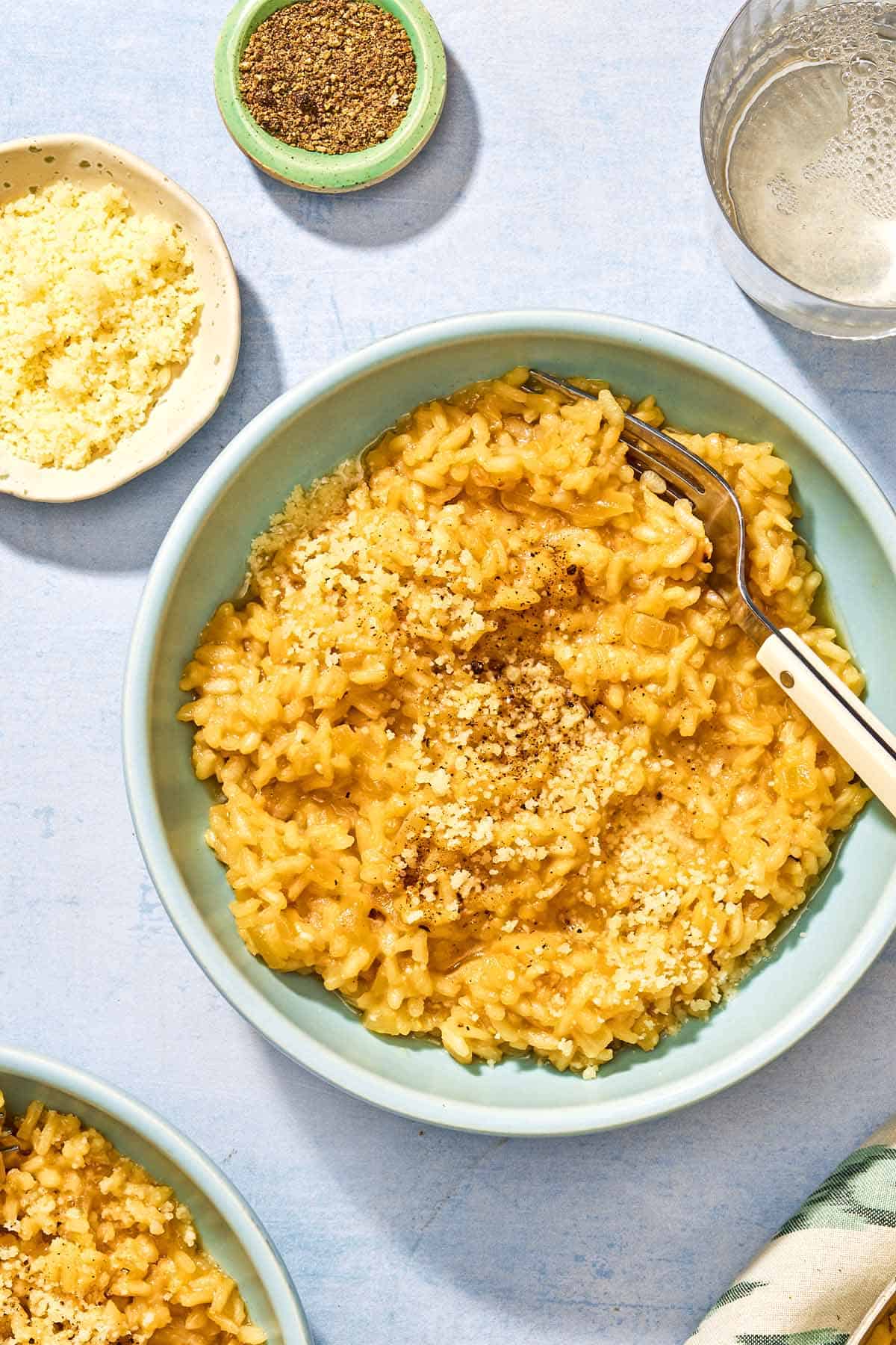 Risotto in a bowl with a fork surrounded by small bowls of Parmigiano-Reggiano cheese and black pepper and a glass of white wine.