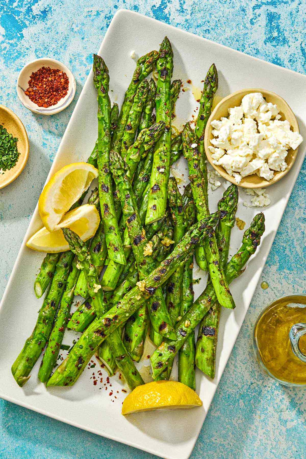 Grilled asparagus on a platter with lemon wedges and a bowl of crumbled feta. Next to this is a bottle of olive oil, and bowls of aleppo pepper and chopped mint.