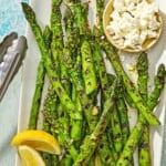 Grilled asparagus on a platter with lemon wedges, a bowl of crumbled feta, and a pair of tongs.