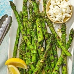 Grilled asparagus on a platter with lemon wedges, a bowl of crumbled feta, and a pair of tongs.