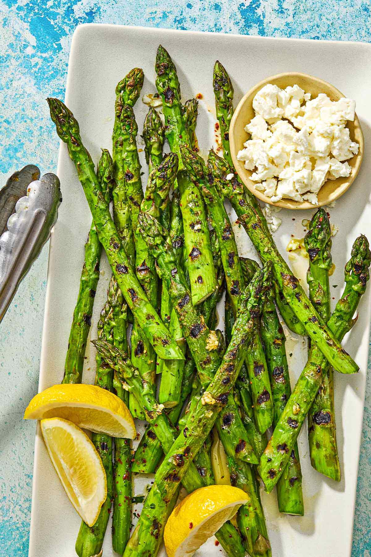 Grilled asparagus on a platter with lemon wedges, a bowl of crumbled feta, and a pair of tongs.