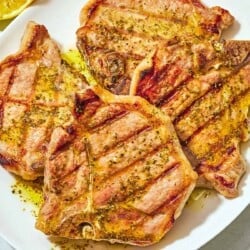 Four grilled pork chops topped with ladorigano dressing on a plate next to 2 juiced lemon halves.
