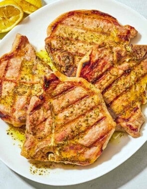 Four grilled pork chops topped with ladorigano dressing on a plate next to 2 juiced lemon halves.
