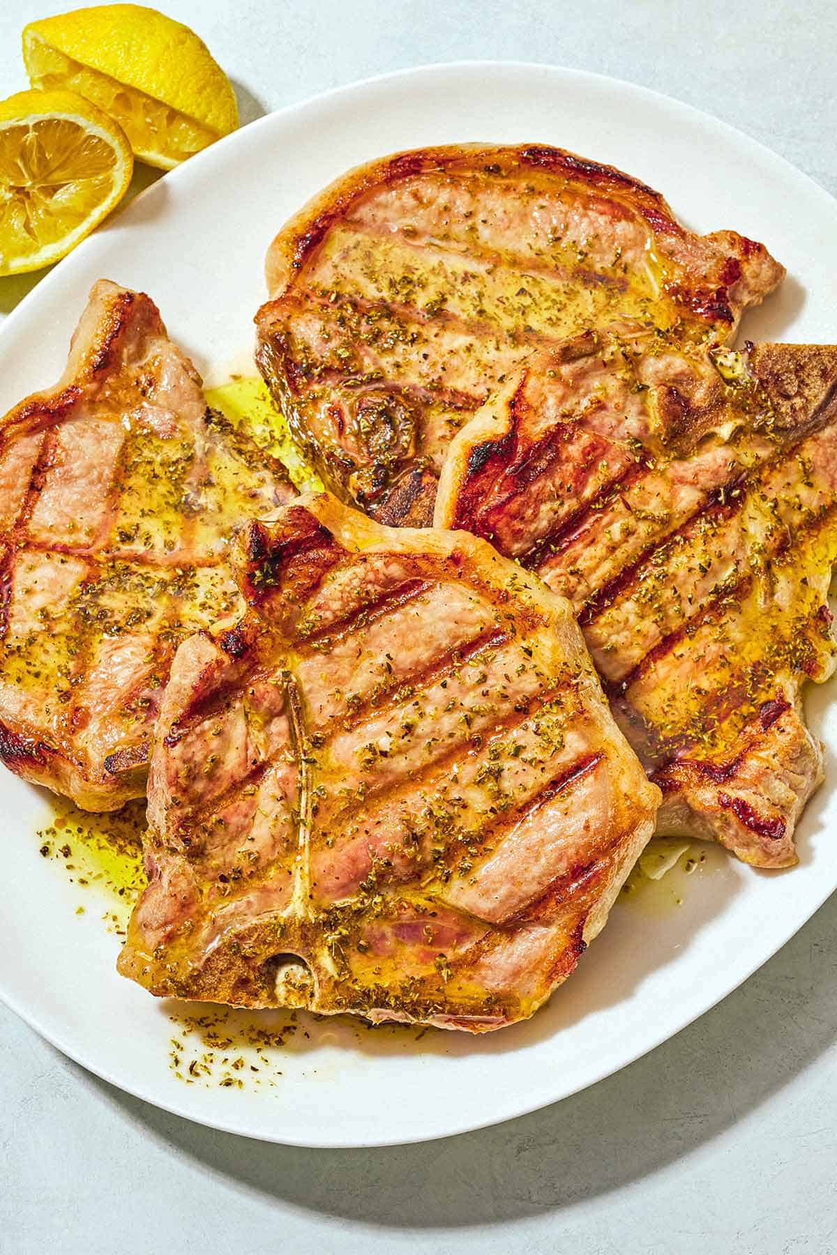 Four grilled pork chops topped with ladorigano dressing on a plate next to 2 juiced lemon halves.
