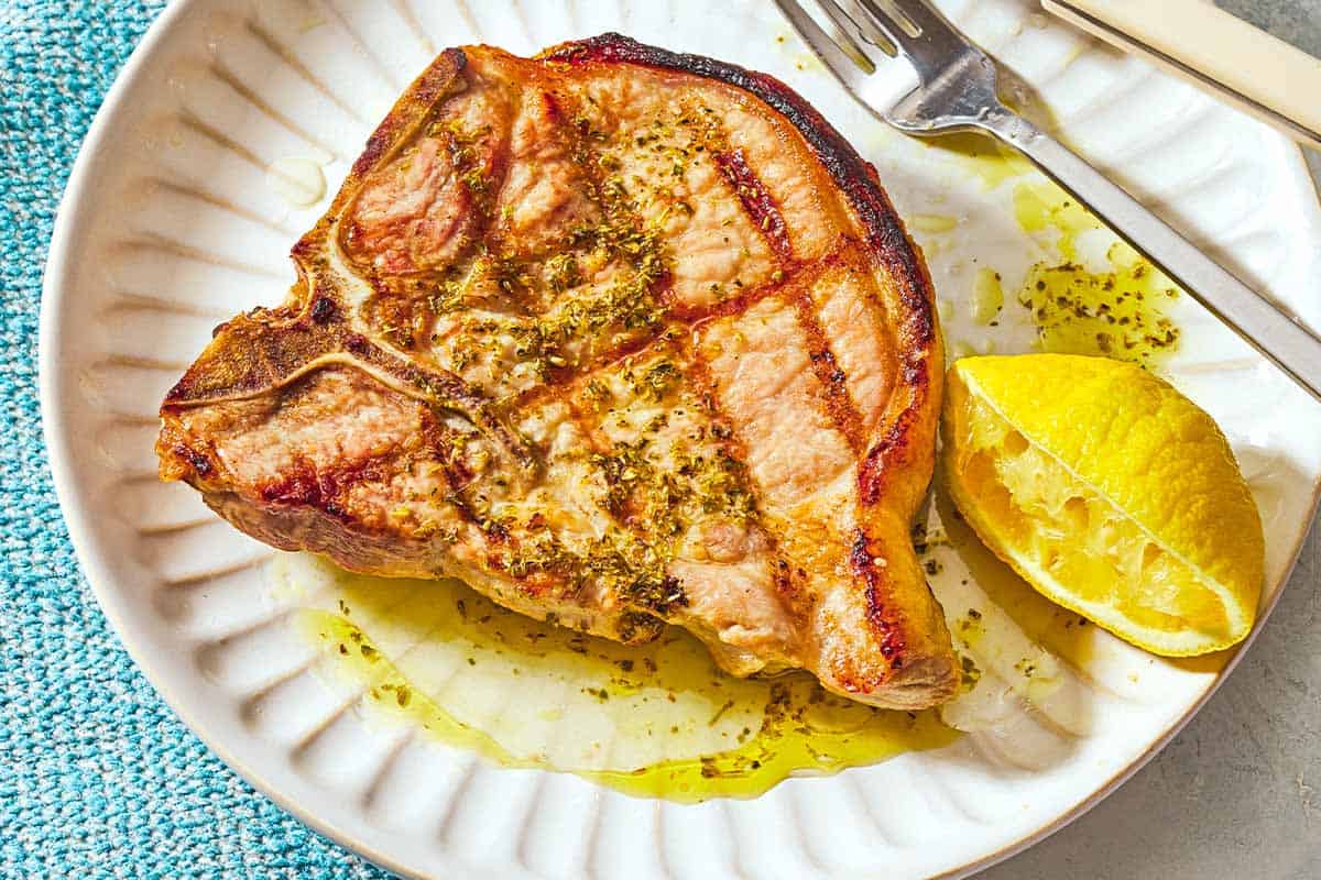 A grilled pork chop topped with ladorigano dressing on a plate with a fork and a juiced lemon half.