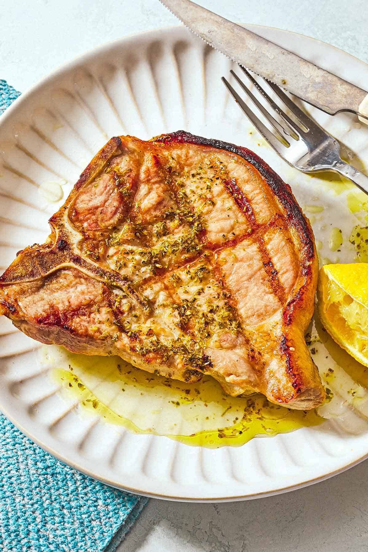A grilled pork chop topped with ladorigano dressing on a plate with a fork, knife, and a juiced lemon half.
