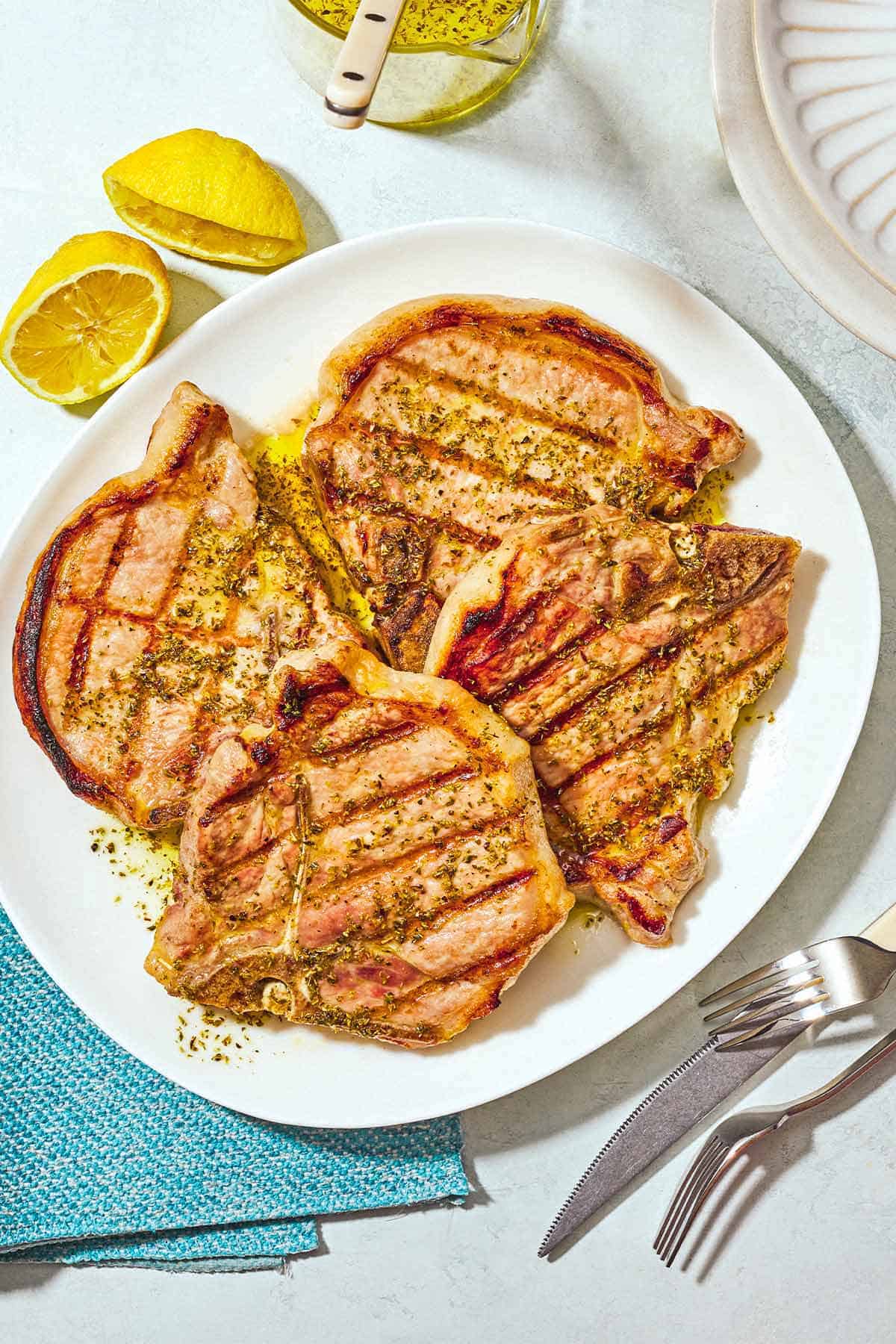 Four grilled pork chops topped with ladorigano dressing on a plate surrounded by 2 forks, a knife, a cloth napkin, and 2 juiced lemon halves.