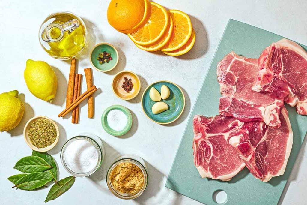 Ingredients for grilled pork chops including bone in pork chops, salt, brown sugar, garlic, orange, cinnamon ticks, whole cloves, whole peppercorns, bay leaves, lemons, olive oil, and dried oregano.