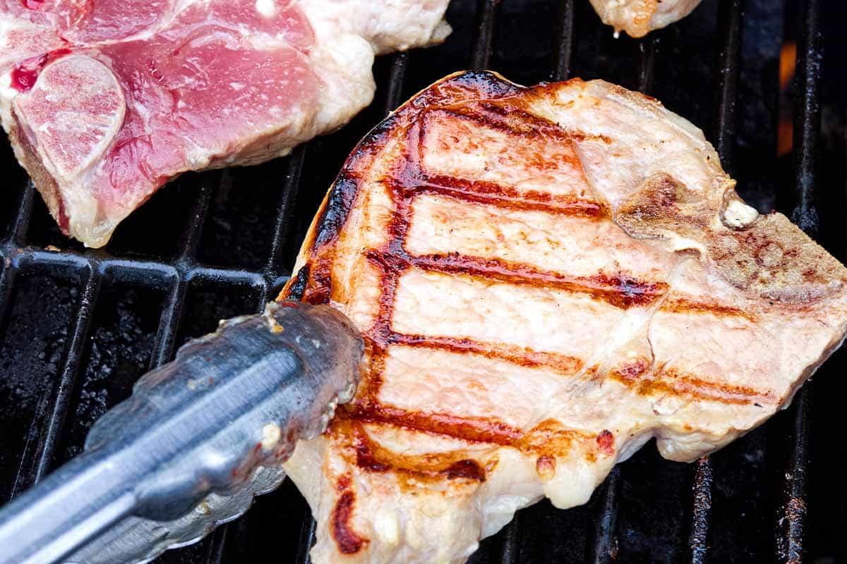 Two pork chops cooking on a grill, one was just turned using a pair of tongs.