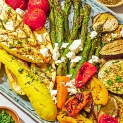 A variety of grilled vegetables on a platter topped with crumbled feta, parsley, basil, za'atar and sumac. Next to this is some basil and small bowls of chopped parsley, sumac and za'atar.