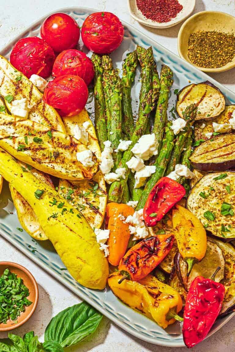 A variety of grilled vegetables on a platter topped with crumbled feta, parsley, basil, za'atar and sumac. Next to this is some basil and small bowls of chopped parsley, sumac and za'atar.