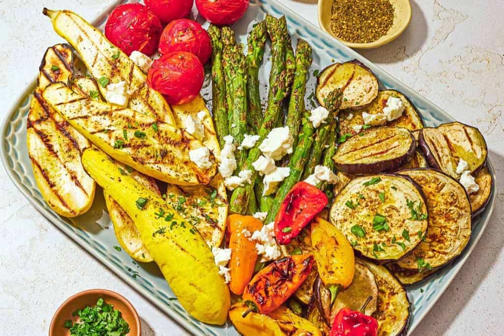 A variety of grilled vegetables on a platter topped with crumbled feta, parsley, basil, za'atar and sumac. Next to this are bowls of za'atar and chopped parsley.