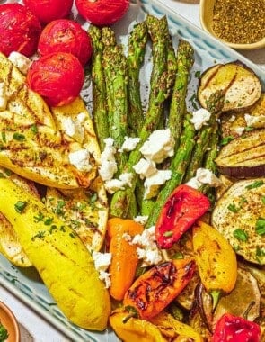 A variety of grilled vegetables on a platter topped with crumbled feta, parsley, basil, za'atar and sumac. Next to this are bowls of za'atar and chopped parsley.