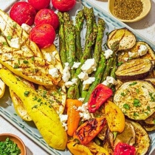 A variety of grilled vegetables on a platter topped with crumbled feta, parsley, basil, za'atar and sumac. Next to this are bowls of za'atar and chopped parsley.