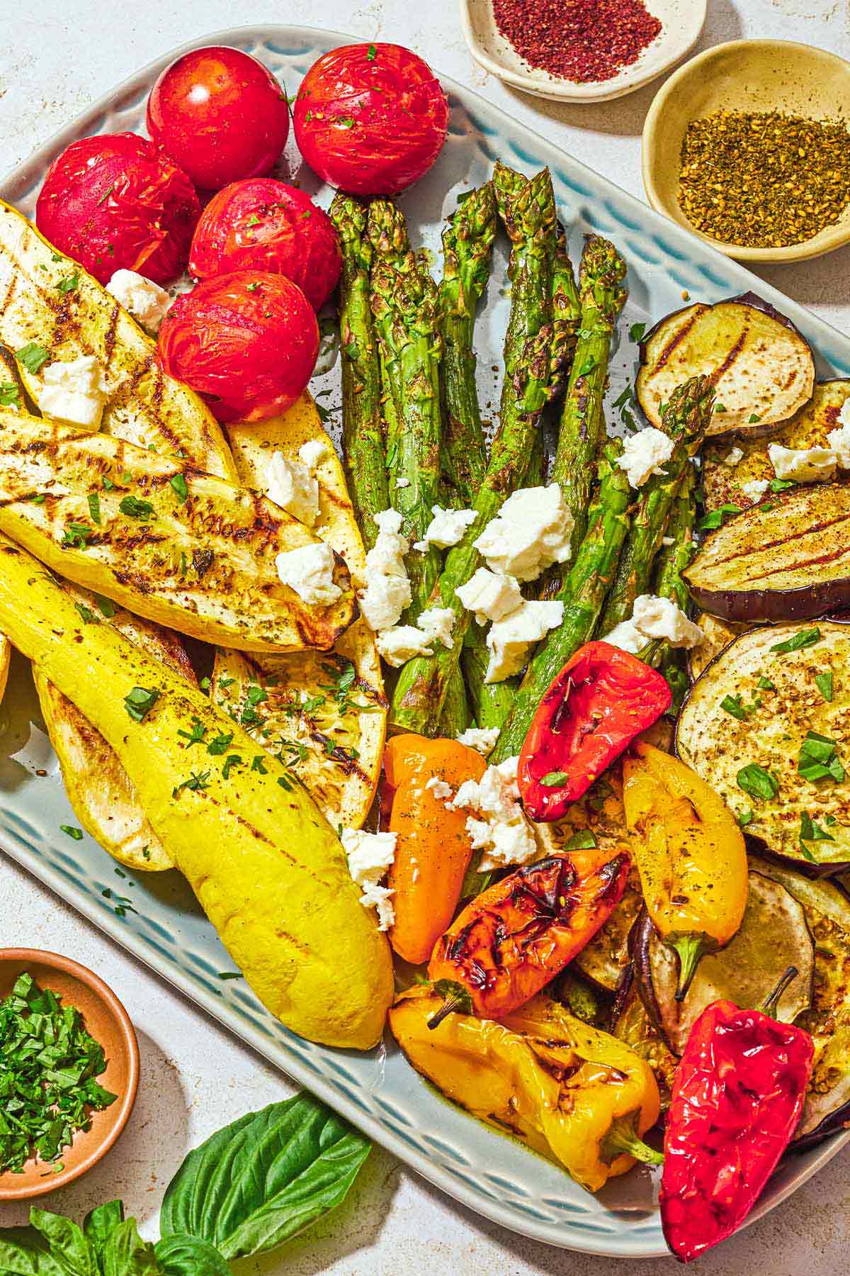 A variety of grilled vegetables on a platter topped with crumbled feta, parsley, basil, za'atar and sumac. Next to this is some basil and small bowls of chopped parsley, sumac and za'atar.