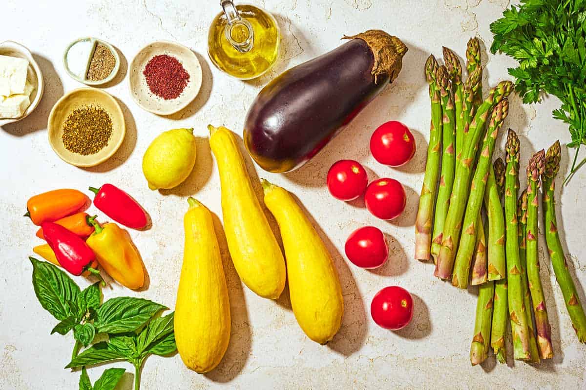 Ingredients for grilled vegetables including parsley, asparagus, tomatoes, eggplant, yellow squash, mini peppers, lemon, basil, olive oil, feta cheese, sumac, za'atar, salt and black pepper.