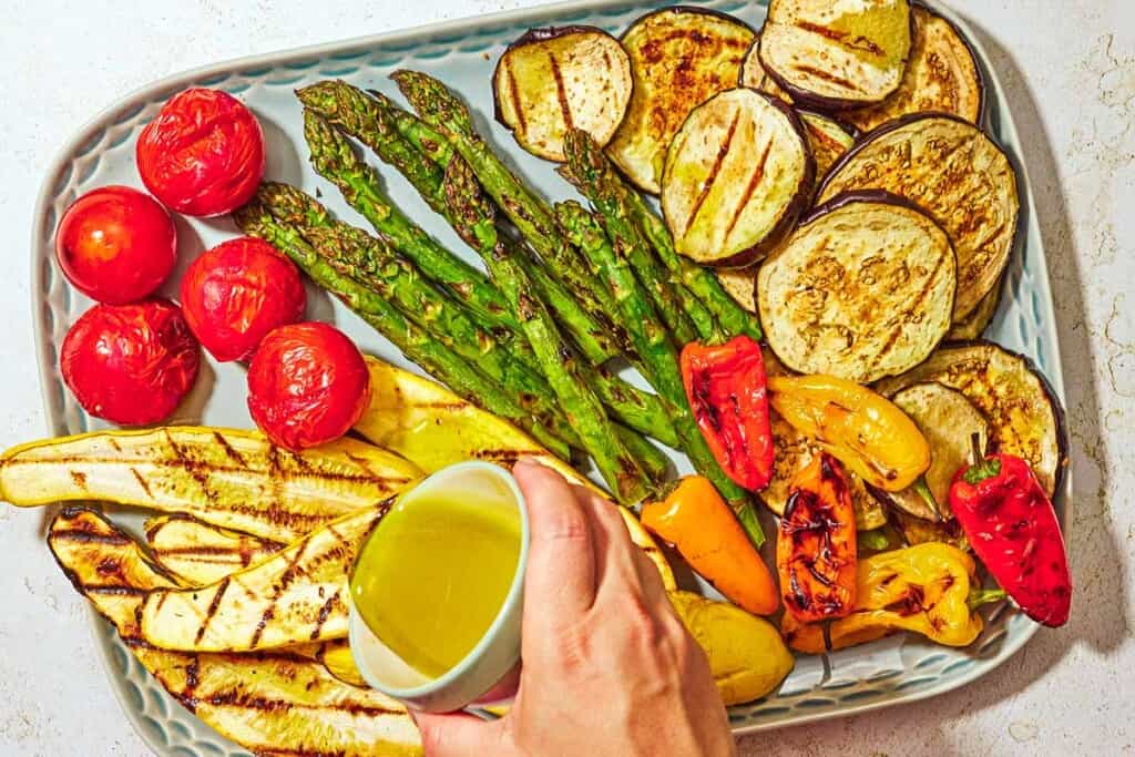 A variety of grilled vegetables on a platter being topped with the lemony dressing.
