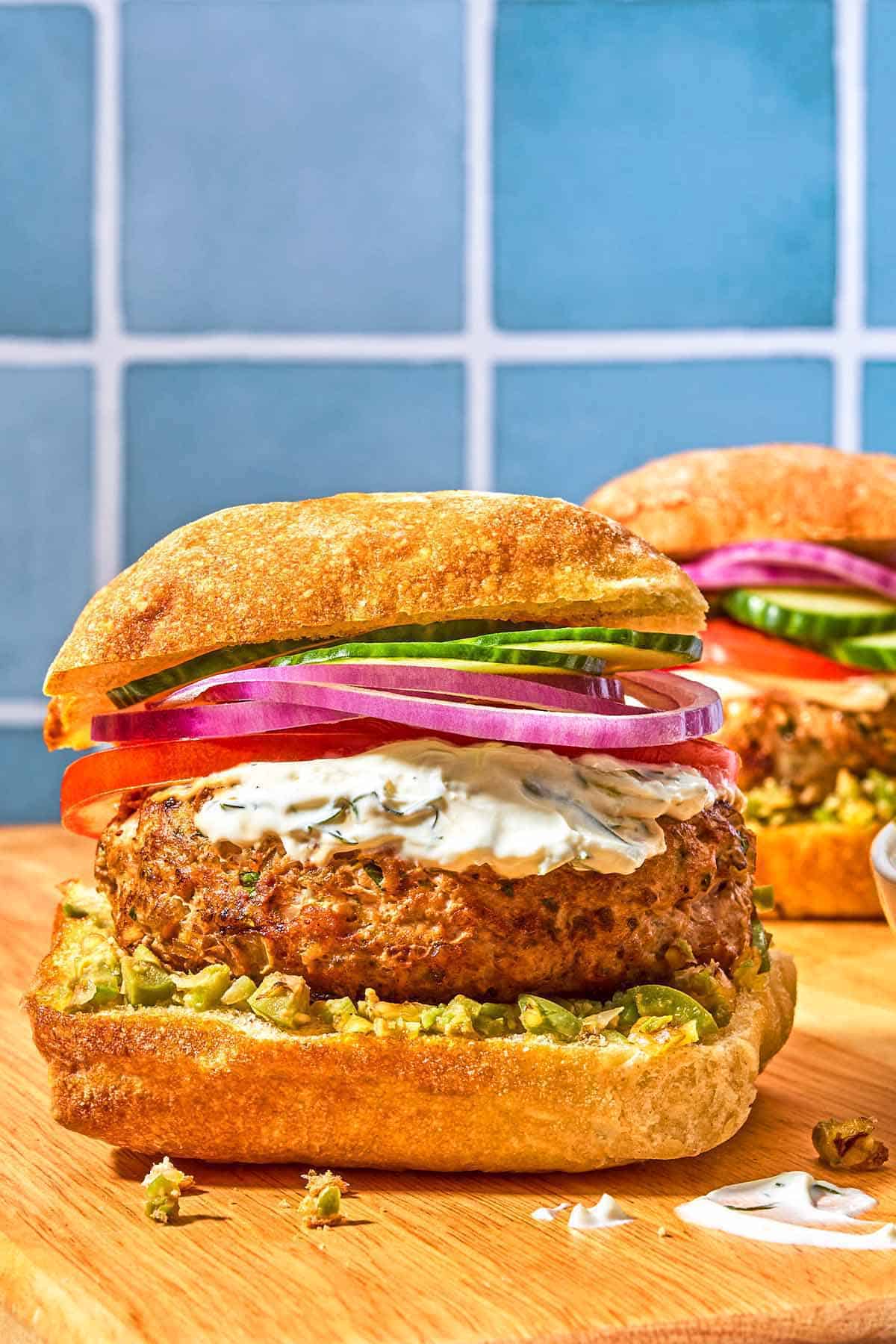 A turkey burger on a ciabatta bun dressed with zeytoon parvardeh, tzatziki, tomatoes, red onions, and cucumbers.