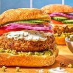 Two turkey burgers on ciabatta rolls dressed with zeytoon parvardeh, tzatziki, tomatoes, red onions, and cucumbers next to a bowl of the zeytoon parvardeh.