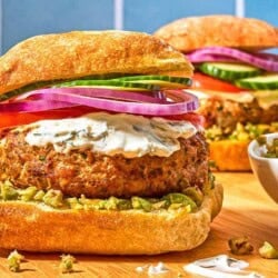 Two turkey burgers on ciabatta rolls dressed with zeytoon parvardeh, tzatziki, tomatoes, red onions, and cucumbers next to a bowl of the zeytoon parvardeh.