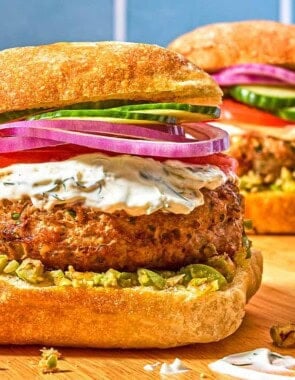 Two turkey burgers on ciabatta rolls dressed with zeytoon parvardeh, tzatziki, tomatoes, red onions, and cucumbers next to a bowl of the zeytoon parvardeh.